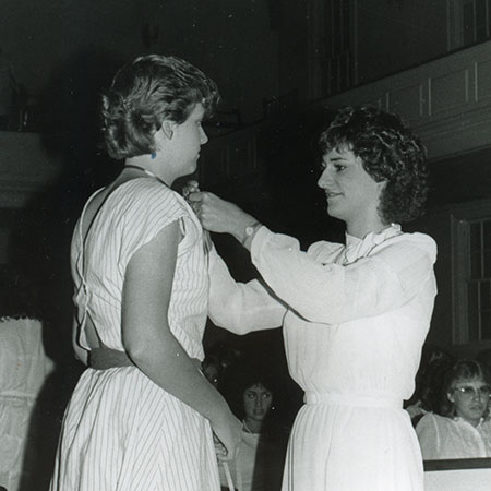 A photo of an upper-class female student tying a ribbon around the neck of a freshman female student.