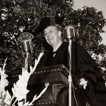 A photo of Eleanor Roosevelt in cap and gown at a lecturn in front of microphones to record her address.